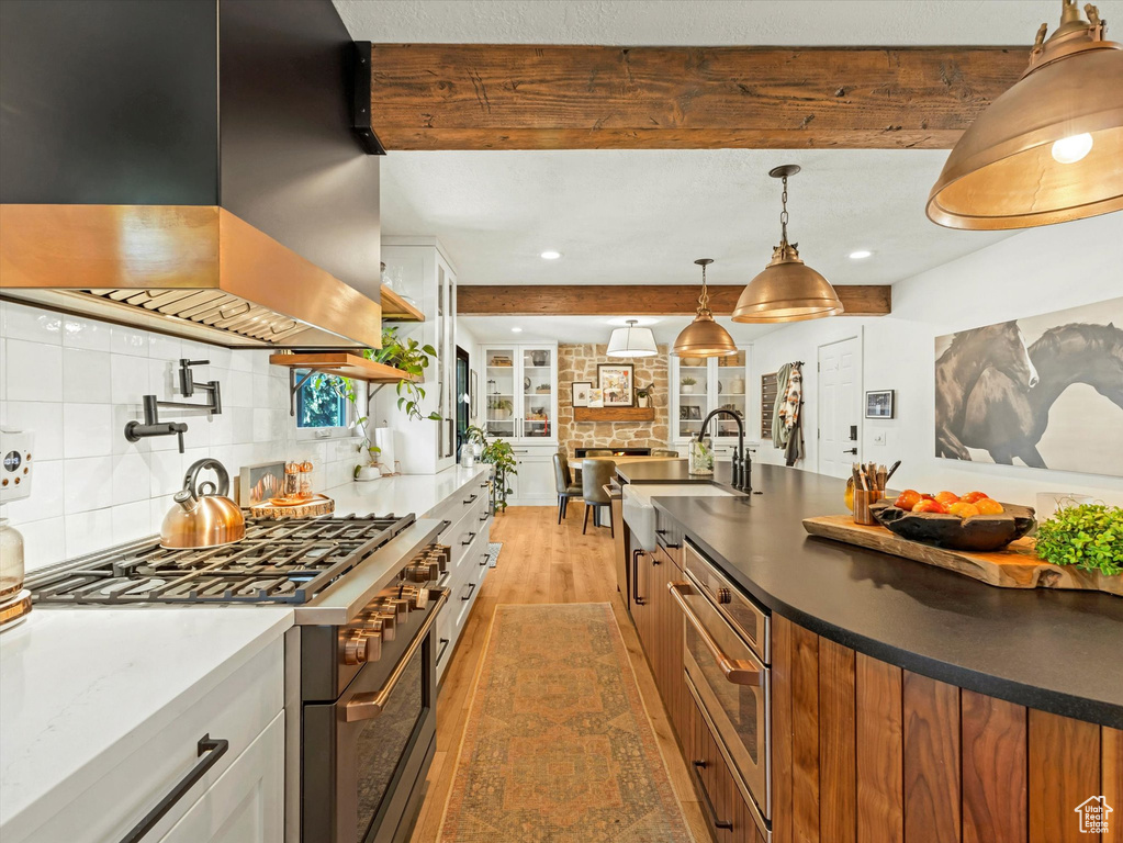 Kitchen with island range hood, appliances with stainless steel finishes, light wood-style floors, beamed ceiling, and tasteful backsplash