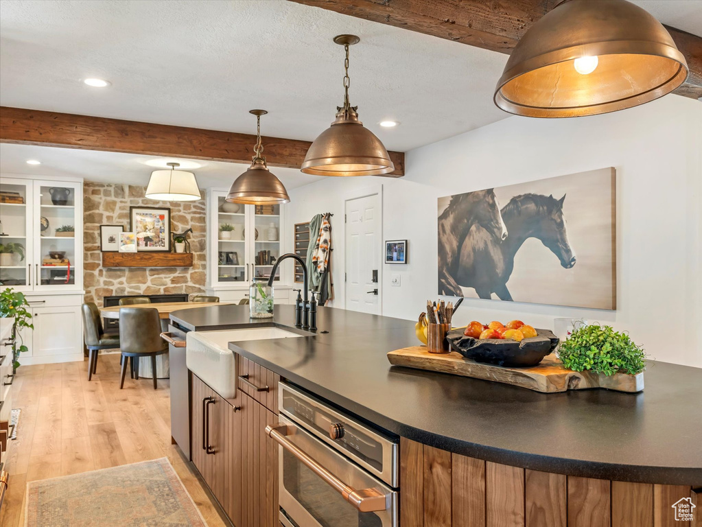 Kitchen with dark countertops, beam ceiling, light wood finished floors, recessed lighting, and an island with sink