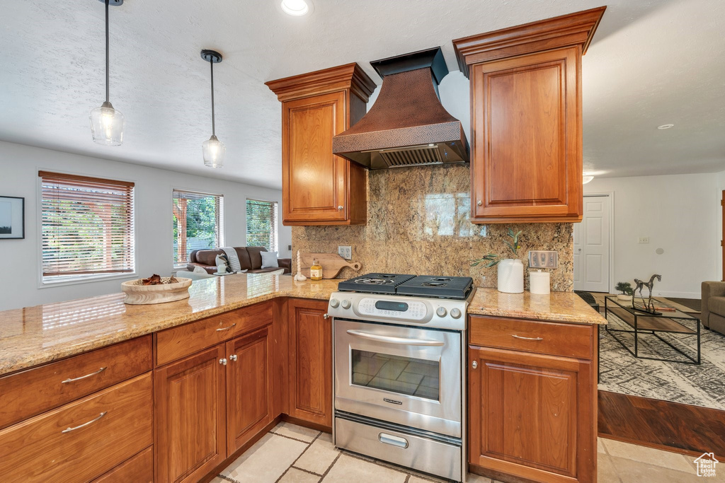 Kitchen featuring custom exhaust hood, stainless steel gas range oven, brown cabinetry, tasteful backsplash, and light stone counters