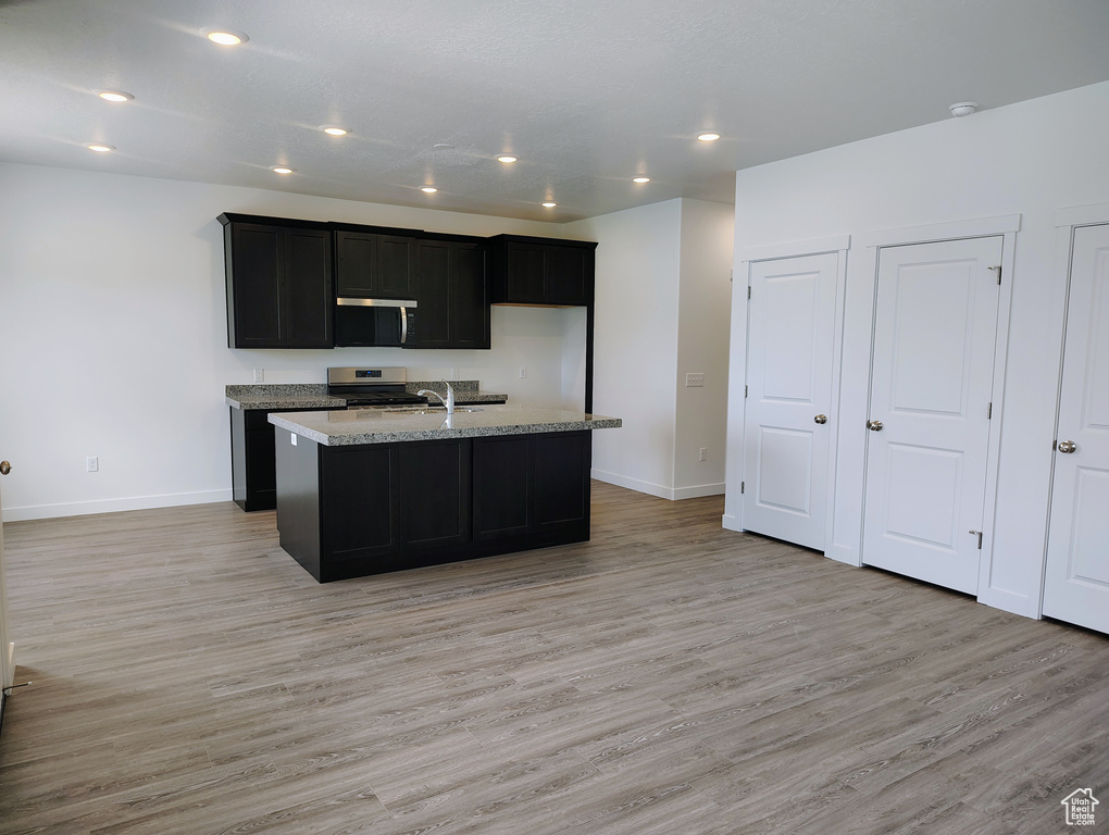 Kitchen with stainless steel appliances, light wood-style floors, dark cabinets, a center island with sink, and light stone counters