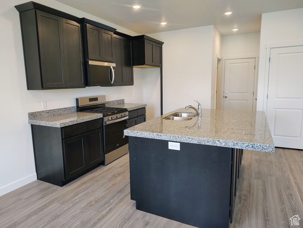 Kitchen with stainless steel appliances, light wood-style flooring, a center island with sink, light stone counters, and recessed lighting