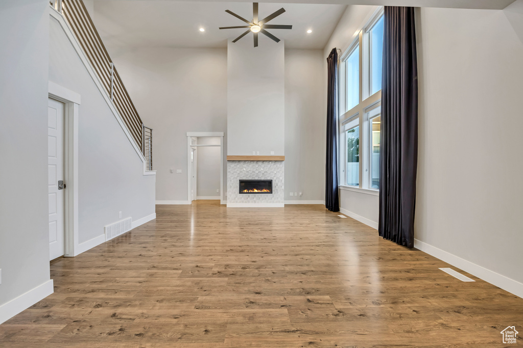 Unfurnished living room featuring a towering ceiling, ceiling fan, healthy amount of natural light, a tiled fireplace, and wood finished floors