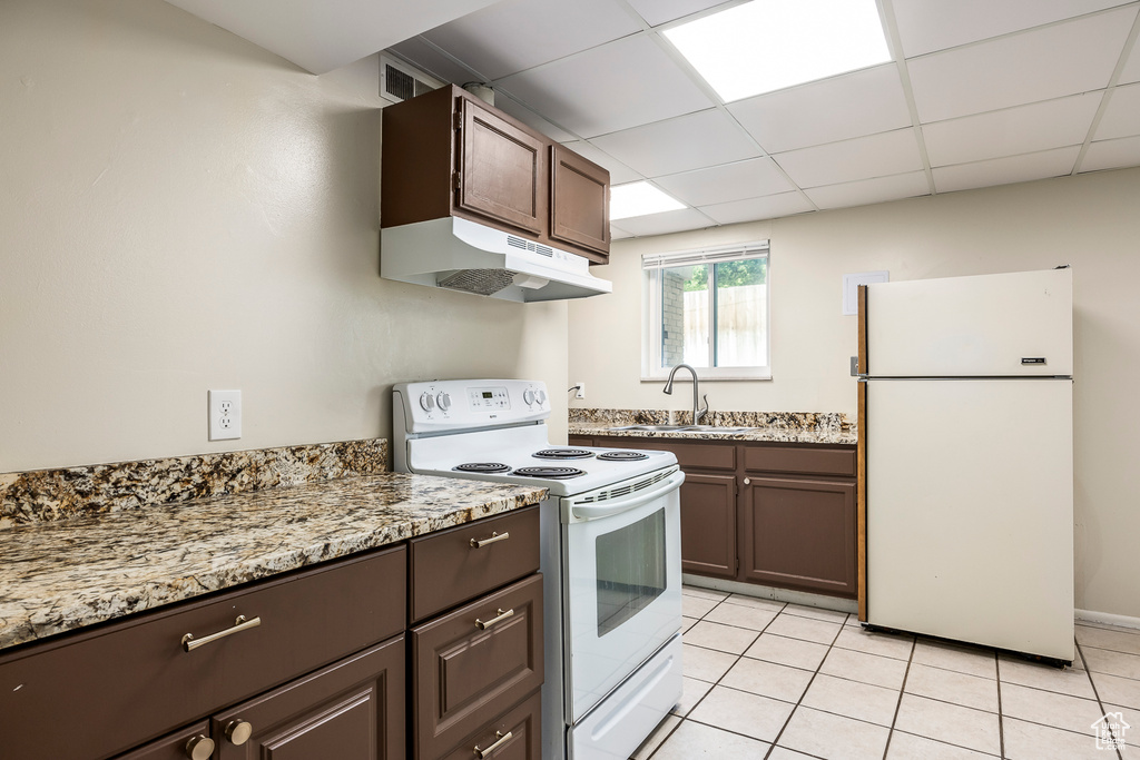 Kitchen with white appliances, under cabinet range hood, light tile patterned floors, dark brown cabinetry, and a drop ceiling