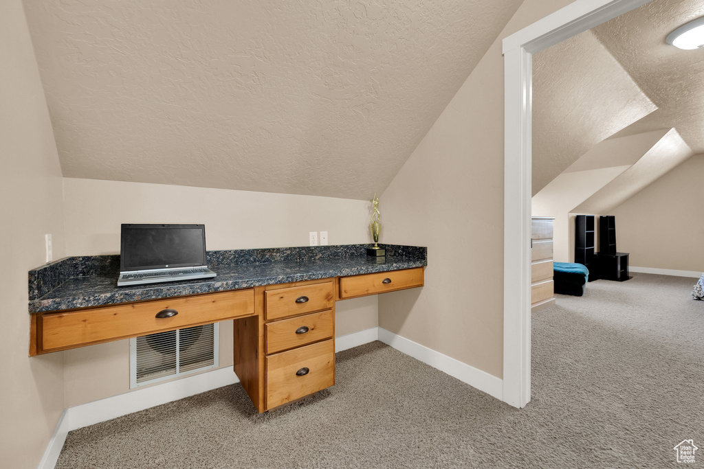Unfurnished office featuring a textured ceiling, vaulted ceiling, built in study area, and light colored carpet