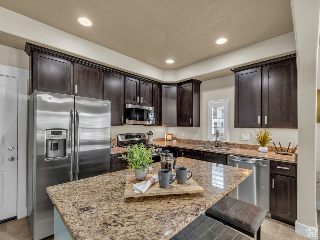 Kitchen featuring appliances with stainless steel finishes, dark brown cabinets, a kitchen breakfast bar, light stone counters, and recessed lighting