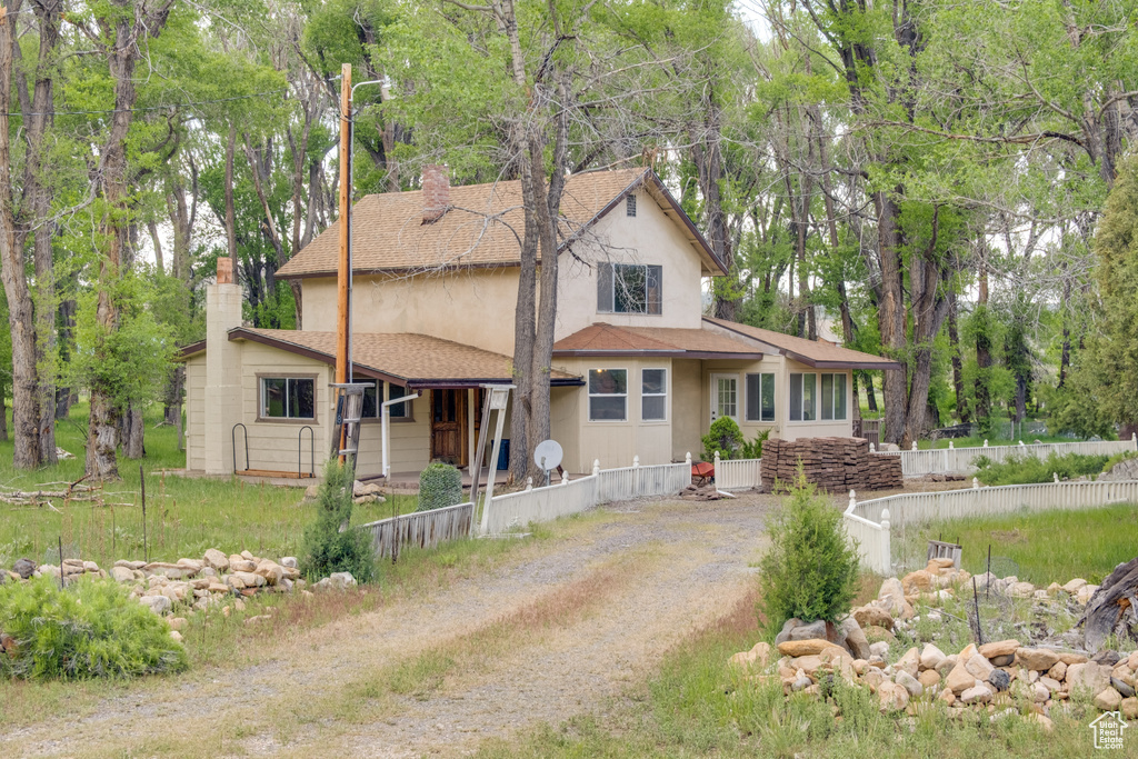View of front of property with a chimney, stucco siding, roof with shingles, dirt driveway, and view of wooded area