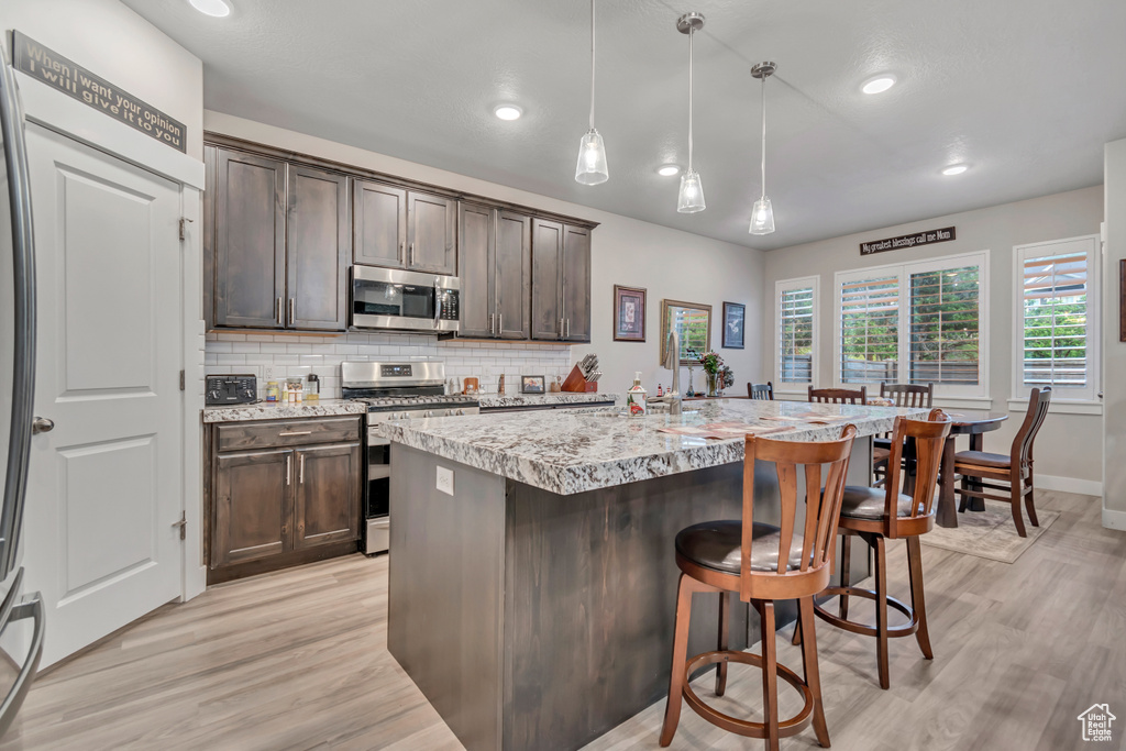 Kitchen with stainless steel appliances, dark brown cabinetry, light wood-style flooring, backsplash, and a breakfast bar