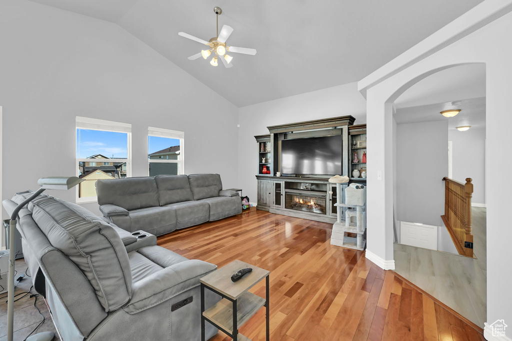 Living area with a ceiling fan, arched walkways, high vaulted ceiling, light wood-style floors, and a glass covered fireplace