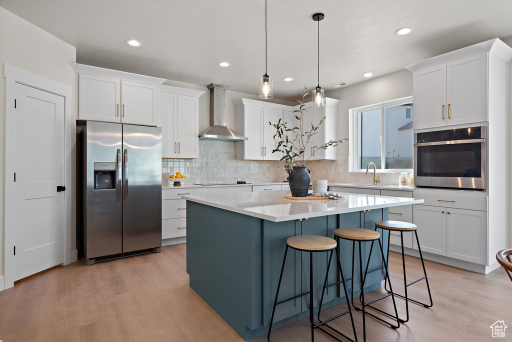 Kitchen featuring stainless steel appliances, wall chimney exhaust hood, light wood-style flooring, white cabinetry, and tasteful backsplash