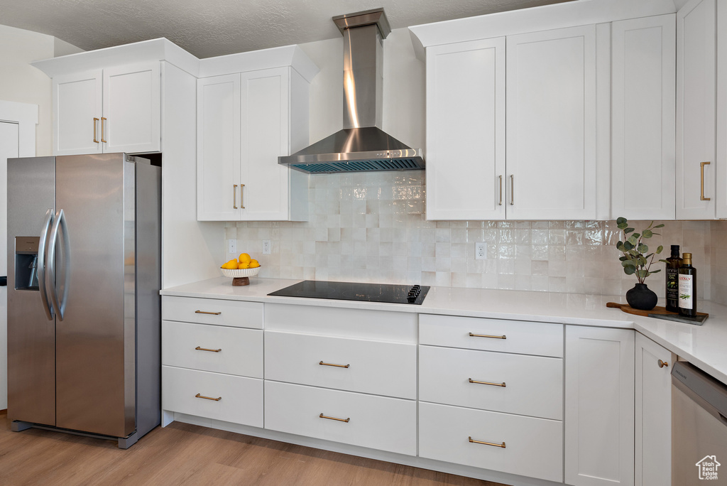 Kitchen featuring appliances with stainless steel finishes, wall chimney range hood, light wood-type flooring, light countertops, and white cabinetry