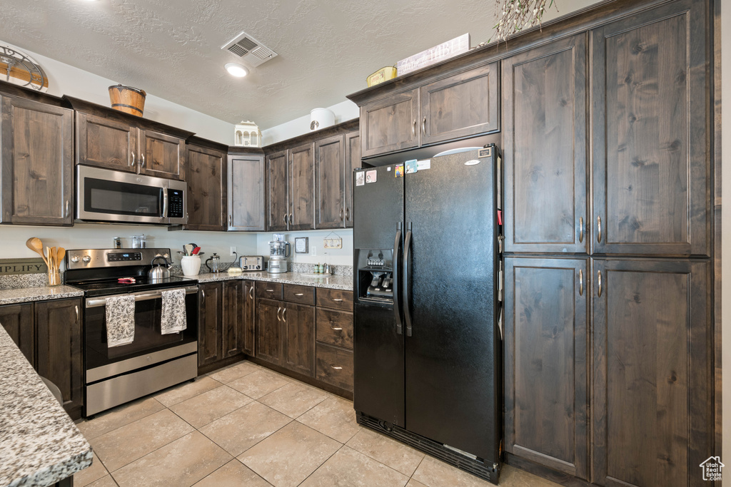 Kitchen featuring stainless steel appliances, dark brown cabinetry, light tile patterned floors, and light stone counters