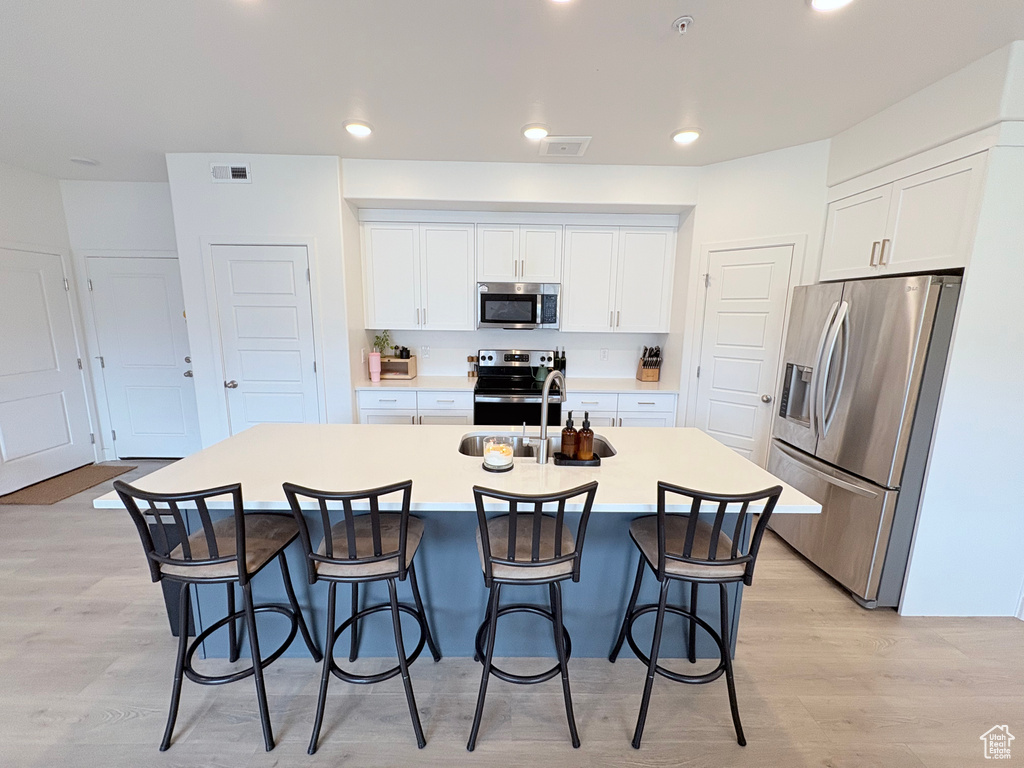 Kitchen with appliances with stainless steel finishes, light wood-type flooring, a kitchen bar, white cabinetry, and a kitchen island with sink