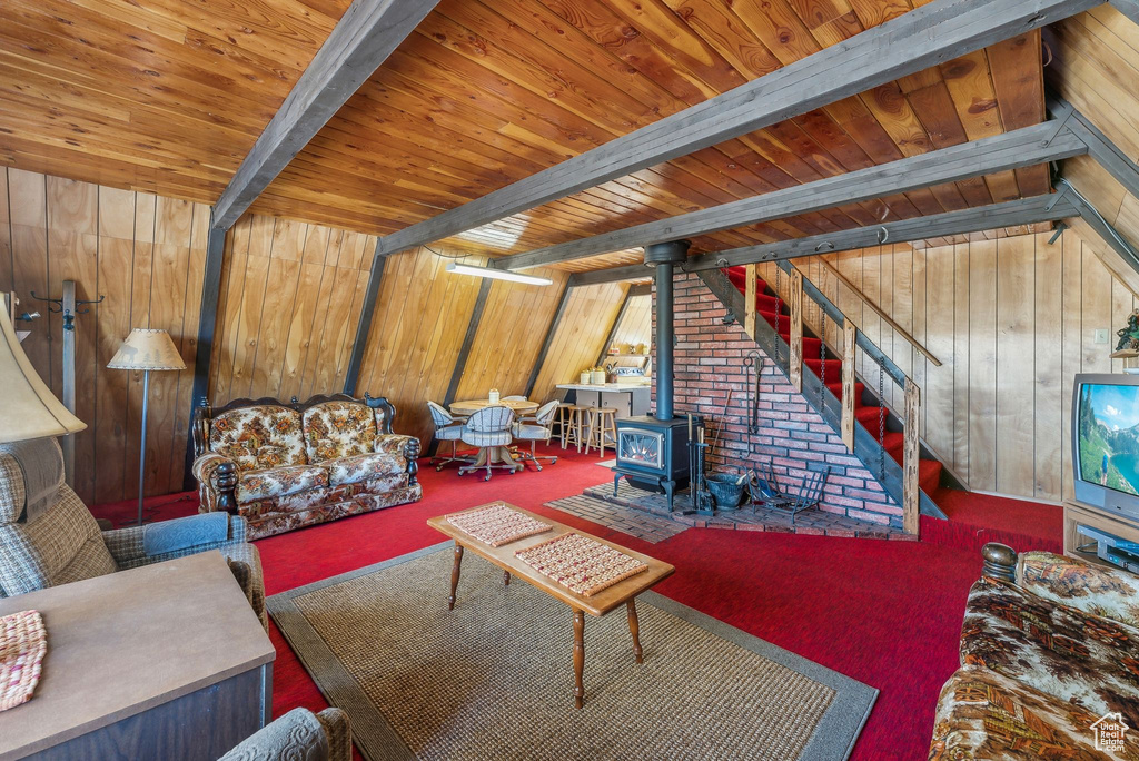 Carpeted living room with a wood stove, wood walls, a wooden ceiling with exposed beams, and stairway