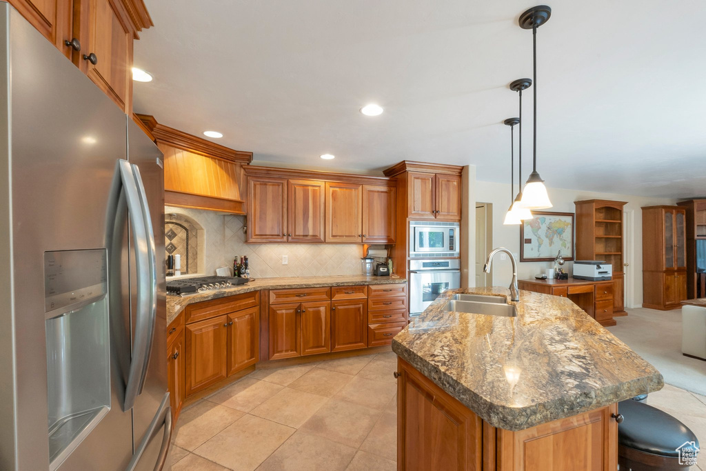 Kitchen featuring stainless steel appliances, brown cabinets, backsplash, a center island with sink, and recessed lighting
