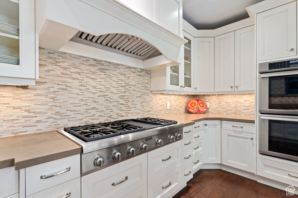 Kitchen featuring stainless steel appliances, custom exhaust hood, white cabinets, glass insert cabinets, and decorative backsplash
