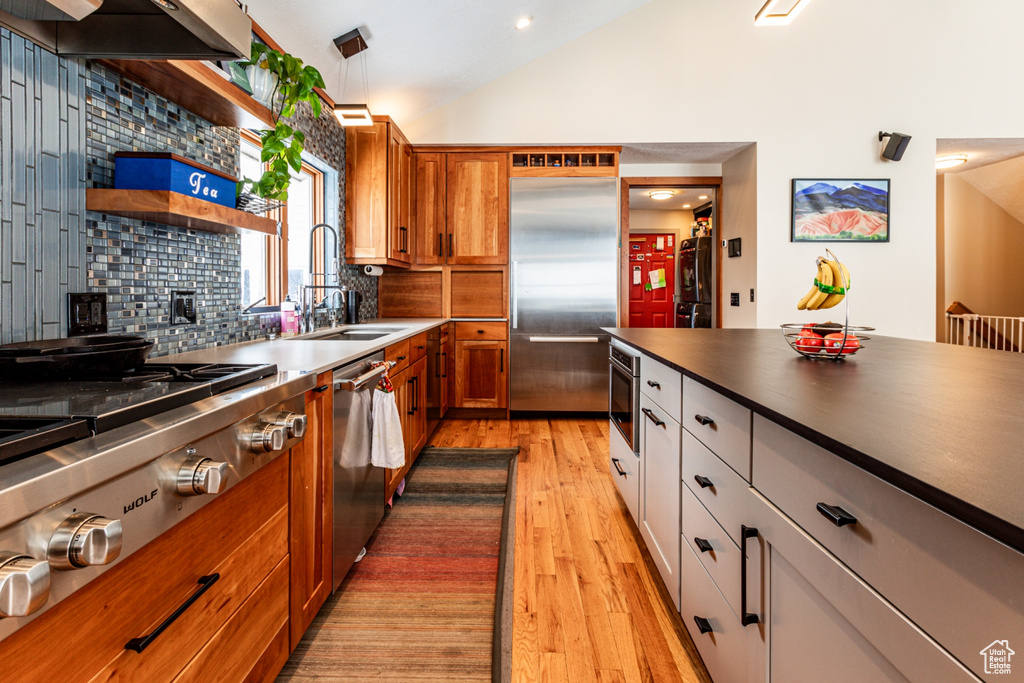 Kitchen with stainless steel appliances, light wood-style flooring, backsplash, brown cabinets, and high vaulted ceiling