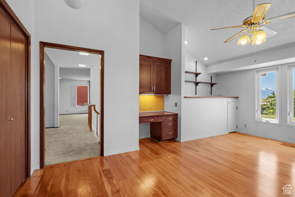 Unfurnished living room featuring built in desk, a textured ceiling, a ceiling fan, light wood-style floors, and high vaulted ceiling
