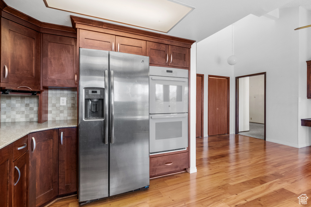 Kitchen with stainless steel fridge, white double oven, tasteful backsplash, light wood-type flooring, and light stone counters