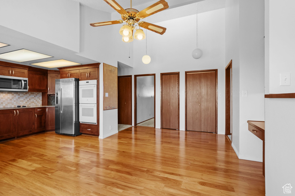Kitchen with appliances with stainless steel finishes, a towering ceiling, a ceiling fan, light wood-style floors, and tasteful backsplash