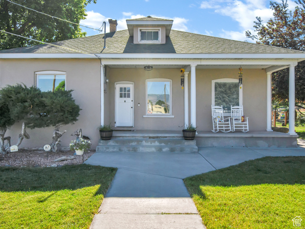 View of front of house featuring covered porch, stucco siding, a chimney, a front lawn, and roof with shingles