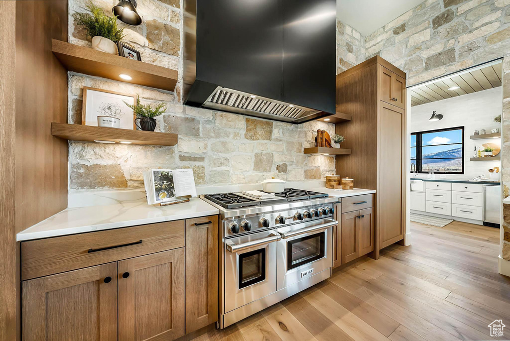 Kitchen with open shelves, wall chimney range hood, range with two ovens, backsplash, and light wood finished floors
