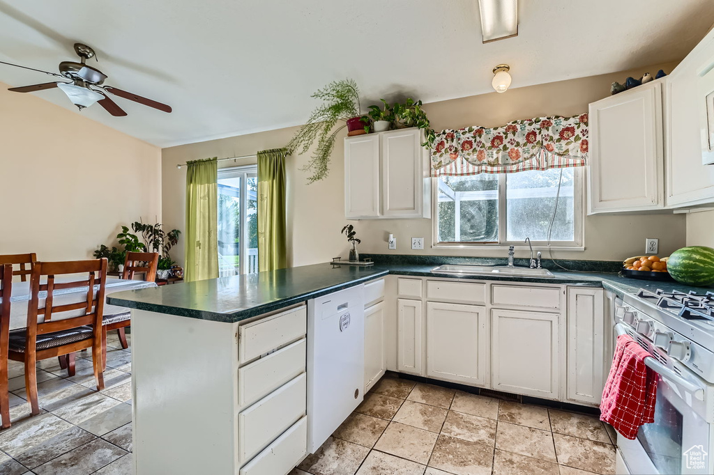 Kitchen with white appliances, a peninsula, ceiling fan, dark countertops, and white cabinetry