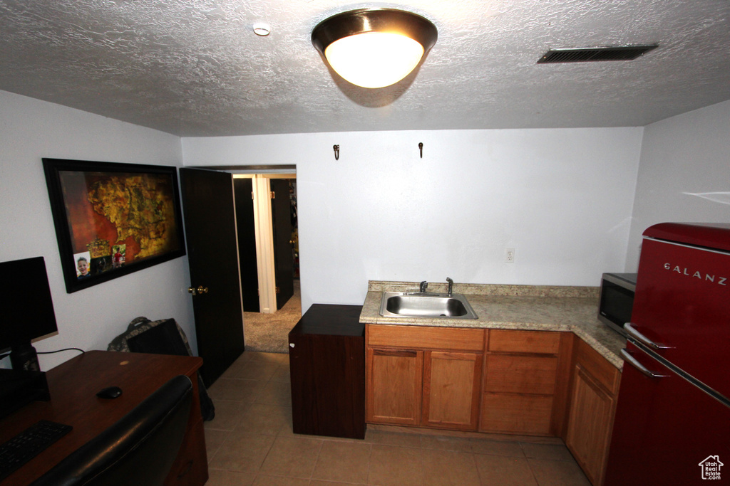 Kitchen featuring freestanding refrigerator, brown cabinets, stainless steel microwave, a textured ceiling, and light tile patterned floors