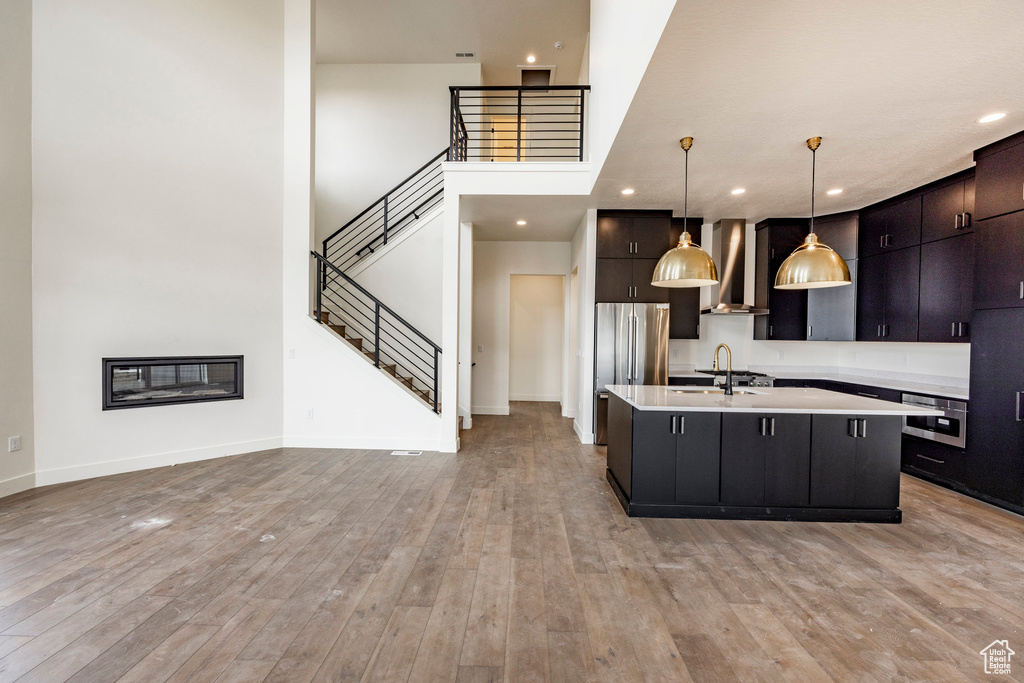 Kitchen featuring wall chimney range hood, light countertops, appliances with stainless steel finishes, a center island with sink, and light wood-type flooring
