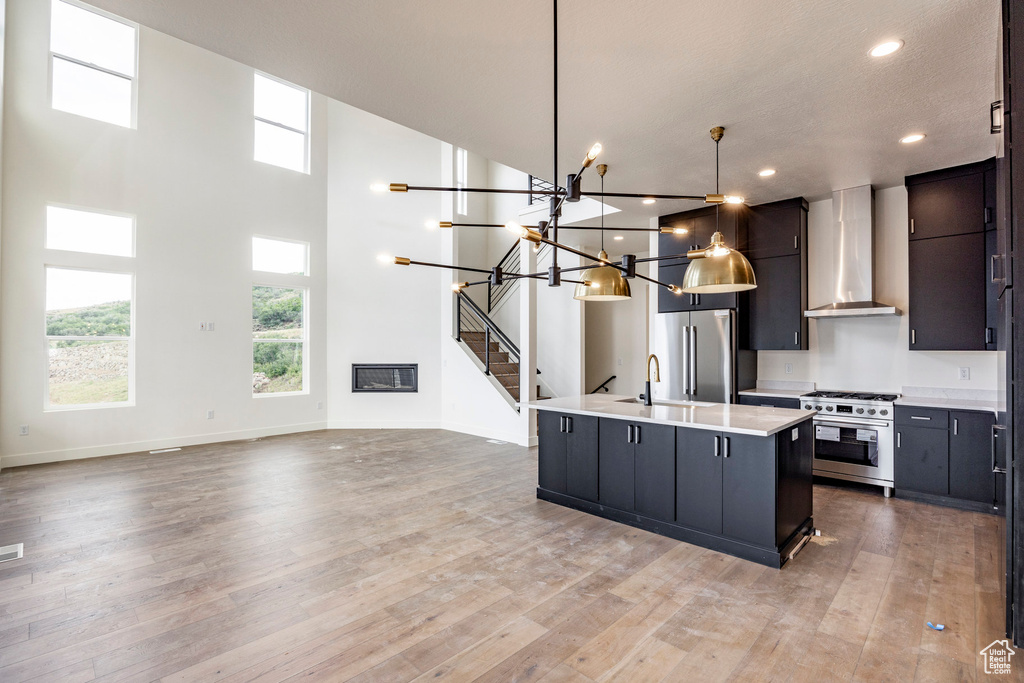 Kitchen featuring stainless steel appliances, wall chimney exhaust hood, light countertops, light wood-type flooring, and a kitchen island with sink