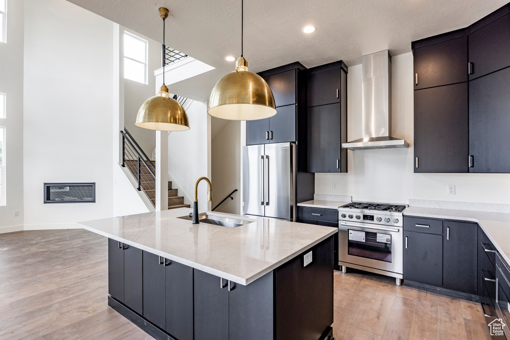 Kitchen with appliances with stainless steel finishes, wall chimney range hood, light wood-style floors, hanging light fixtures, and a center island with sink