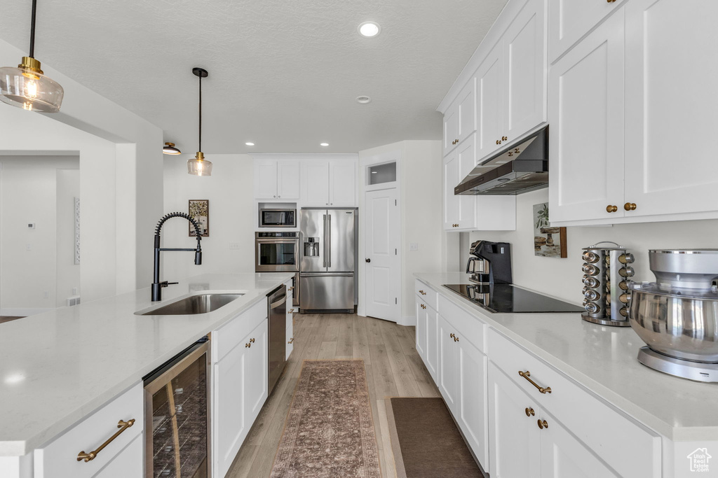 Kitchen with appliances with stainless steel finishes, under cabinet range hood, wine cooler, light wood-style flooring, and white cabinetry