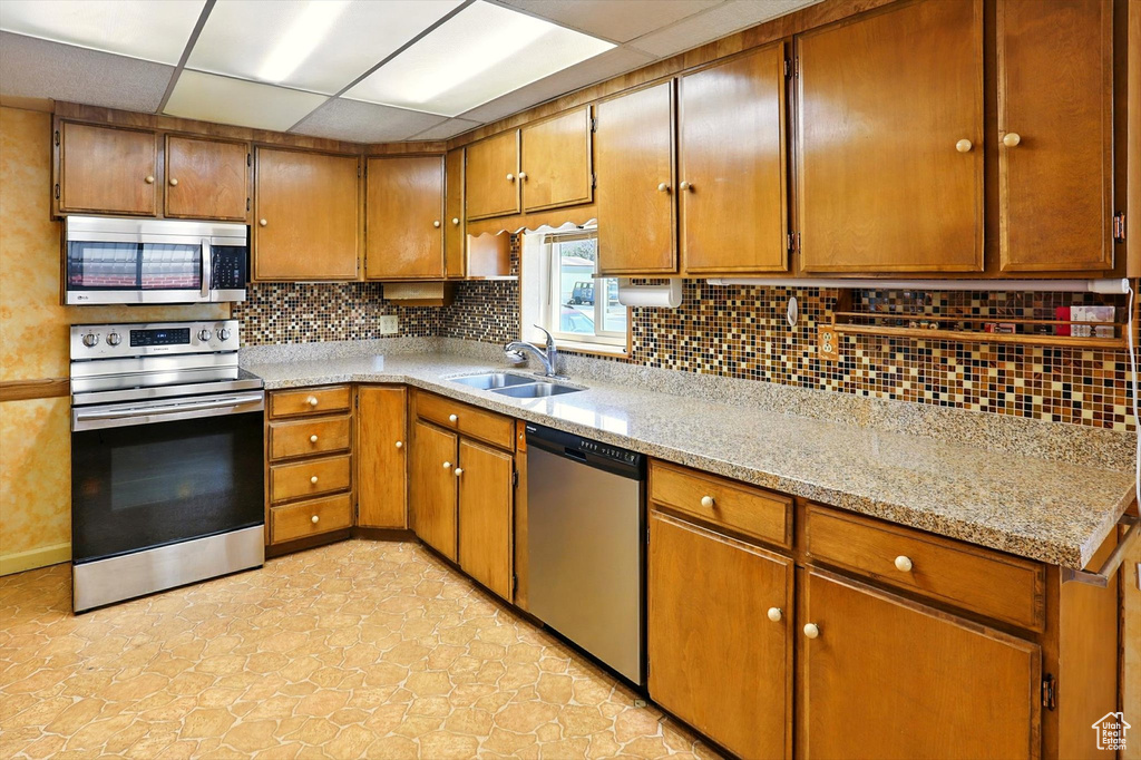 Kitchen featuring appliances with stainless steel finishes, tasteful backsplash, brown cabinetry, a drop ceiling, and light stone countertops
