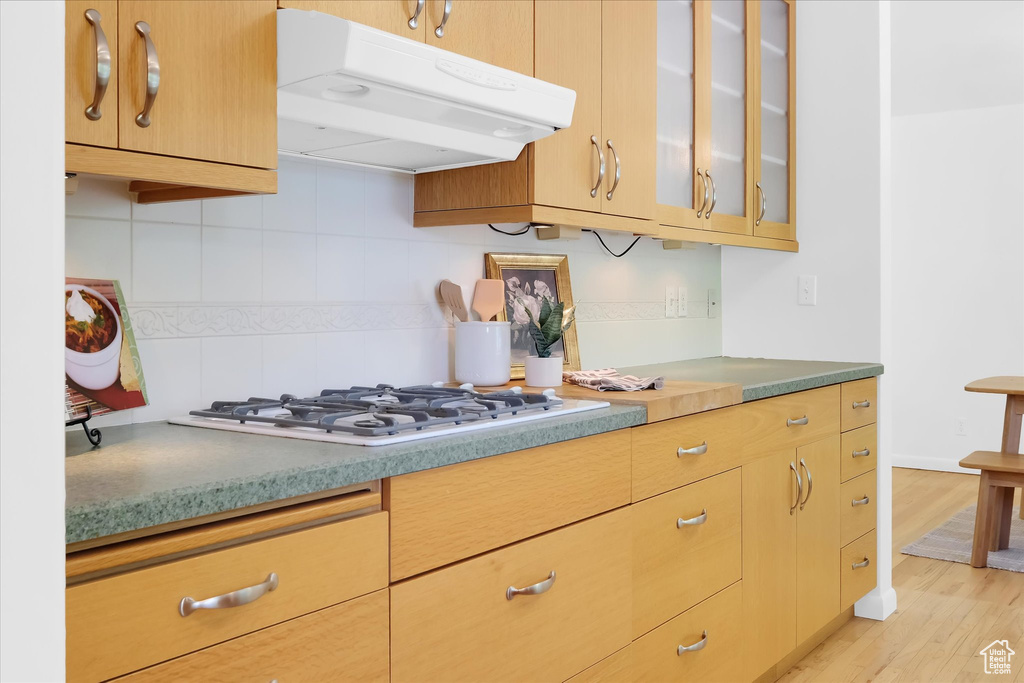 Kitchen with under cabinet range hood, white gas cooktop, decorative backsplash, glass insert cabinets, and light wood-type flooring