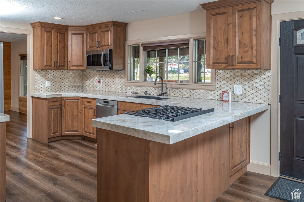 Kitchen featuring appliances with stainless steel finishes, a peninsula, dark wood-style flooring, brown cabinetry, and tasteful backsplash