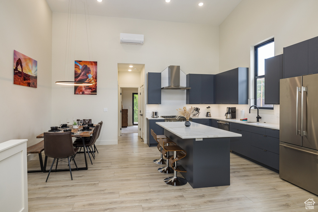 Kitchen featuring appliances with stainless steel finishes, wall chimney exhaust hood, a center island, a high ceiling, and a breakfast bar