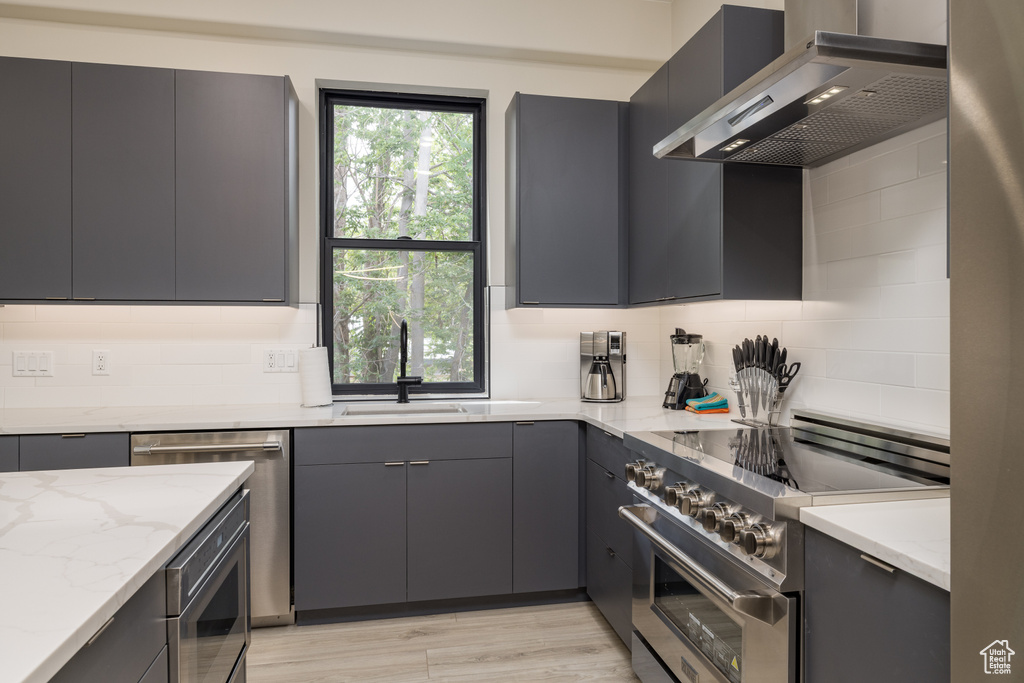 Kitchen featuring wall chimney exhaust hood, appliances with stainless steel finishes, a sink, gray cabinets, and light stone counters