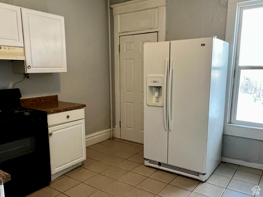 Kitchen with white refrigerator with ice dispenser, black stove, under cabinet range hood, and light tile patterned floors