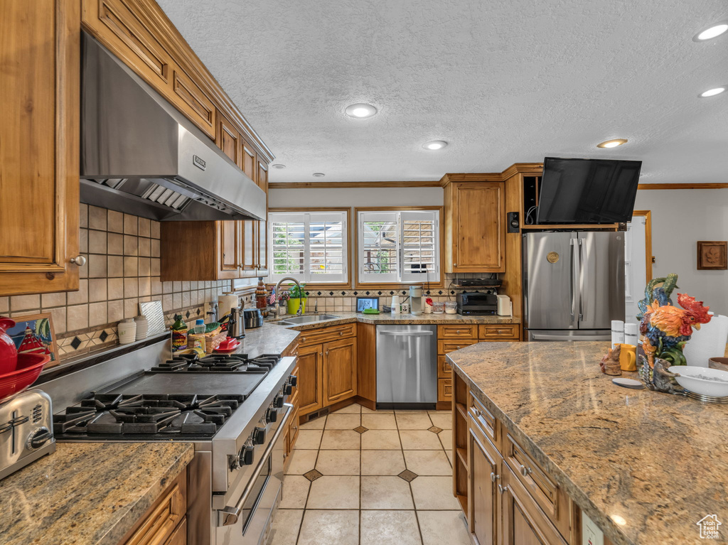 Kitchen with stainless steel appliances, under cabinet range hood, brown cabinetry, ornamental molding, and backsplash