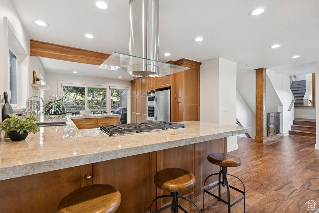 Kitchen featuring appliances with stainless steel finishes, recessed lighting, exhaust hood, brown cabinets, and wood finished floors
