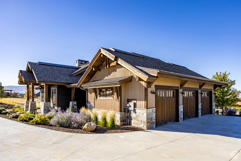 View of property exterior featuring concrete driveway, a shingled roof, roof mounted solar panels, covered porch, and stone siding
