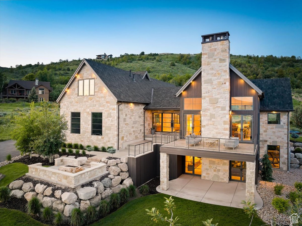 Back of property with stone siding, a balcony, a chimney, and a patio