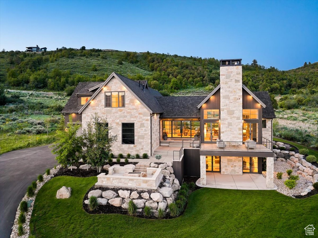 Back of house featuring stone siding, a patio, a yard, a chimney, and a forest view