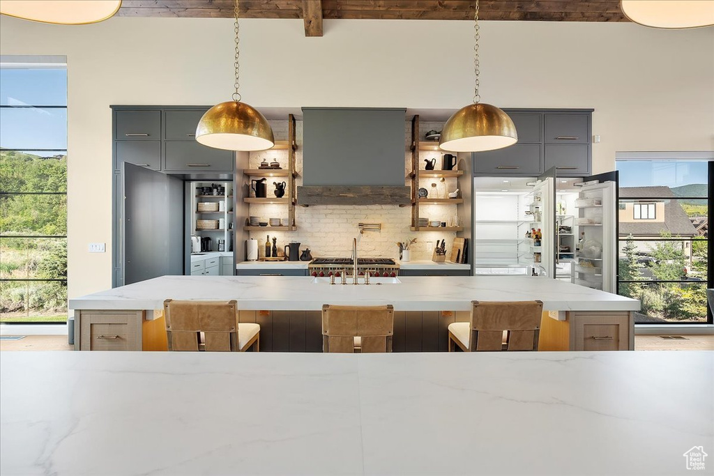 Kitchen with gray cabinetry, backsplash, light stone counters, beamed ceiling, and ventilation hood