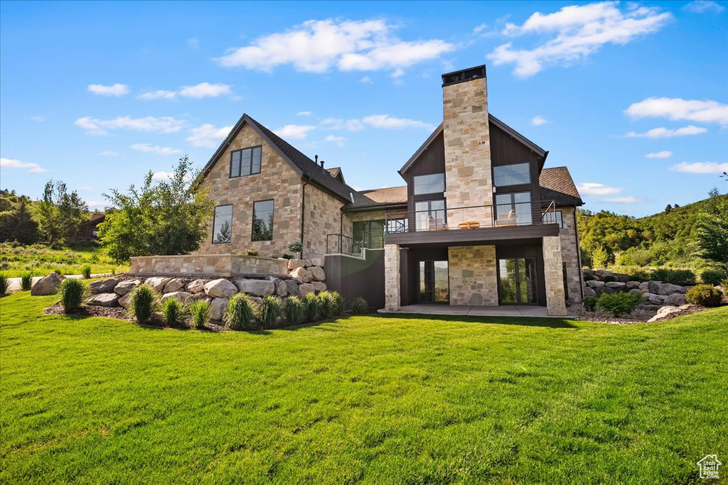 Rear view of property with stone siding, a balcony, a patio area, a yard, and a chimney