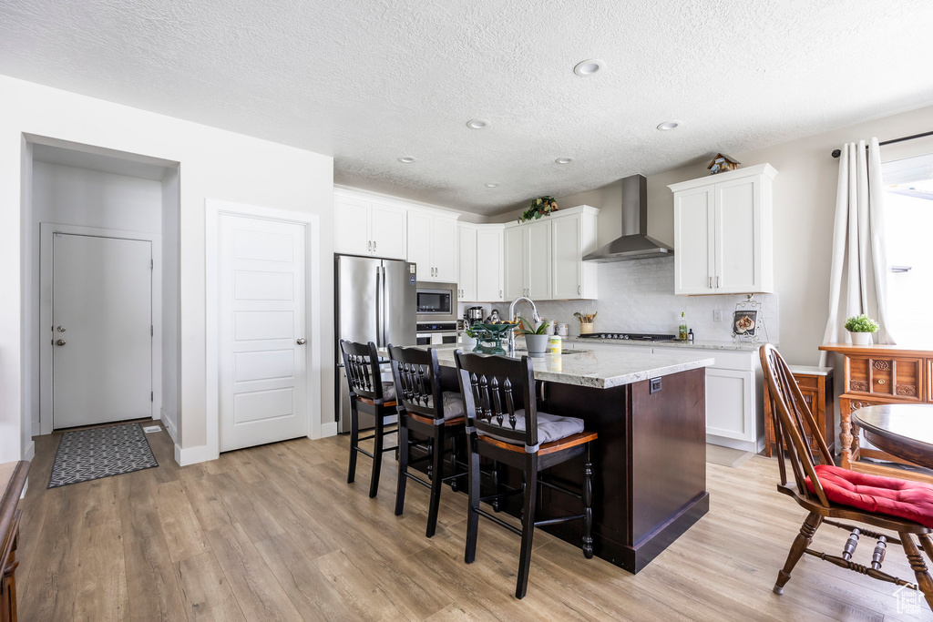 Kitchen with wall chimney range hood, stainless steel appliances, light wood finished floors, a kitchen bar, and an island with sink