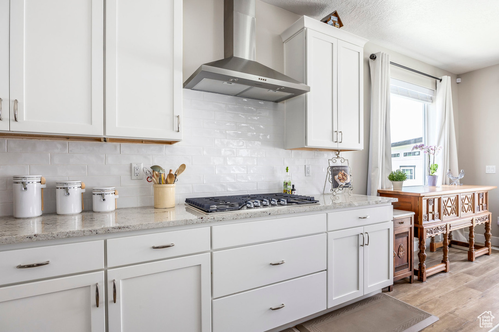 Kitchen featuring wall chimney exhaust hood, stainless steel gas stovetop, light wood-style floors, white cabinets, and decorative backsplash
