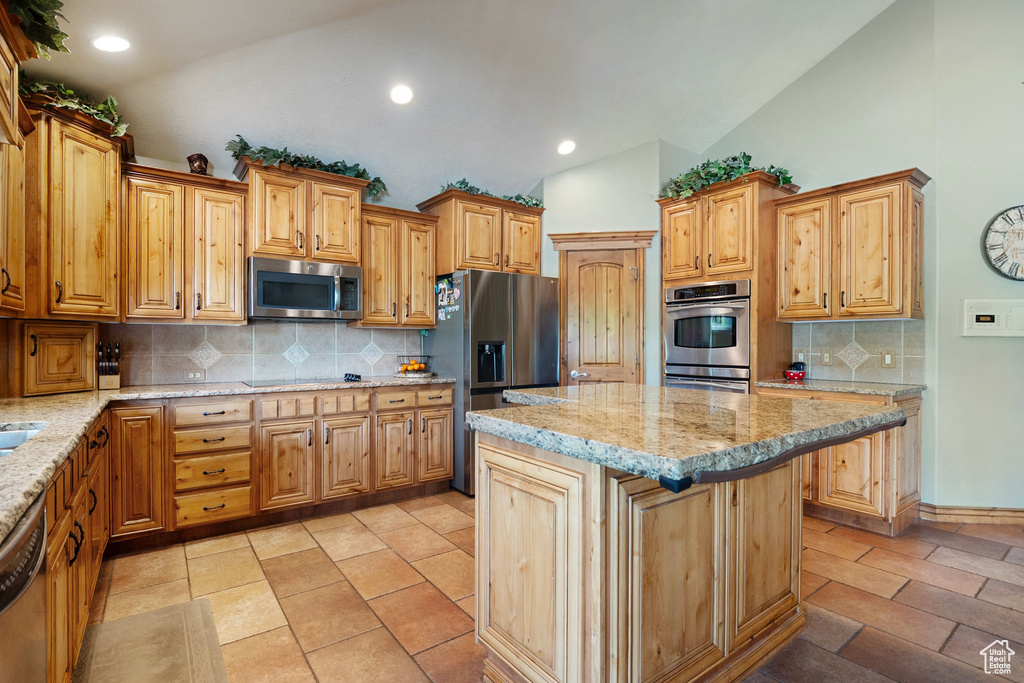 Kitchen featuring stainless steel appliances, lofted ceiling, light stone countertops, decorative backsplash, and a center island