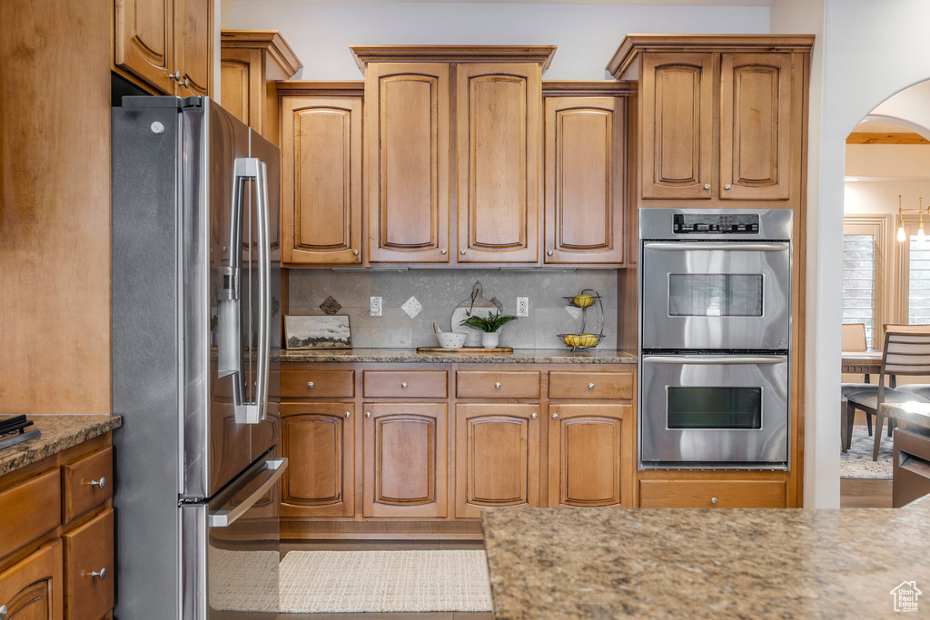 Kitchen featuring appliances with stainless steel finishes, arched walkways, decorative backsplash, light stone counters, and brown cabinetry