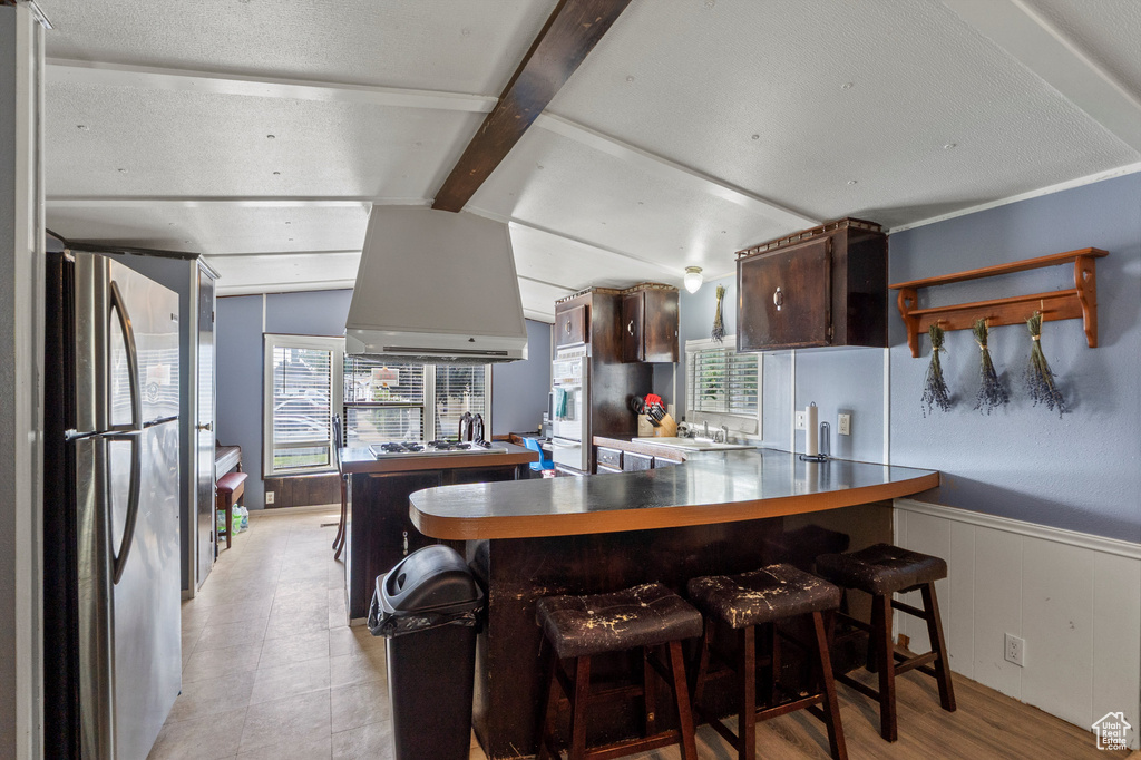 Kitchen featuring freestanding refrigerator, dark brown cabinets, premium range hood, a wainscoted wall, and a peninsula