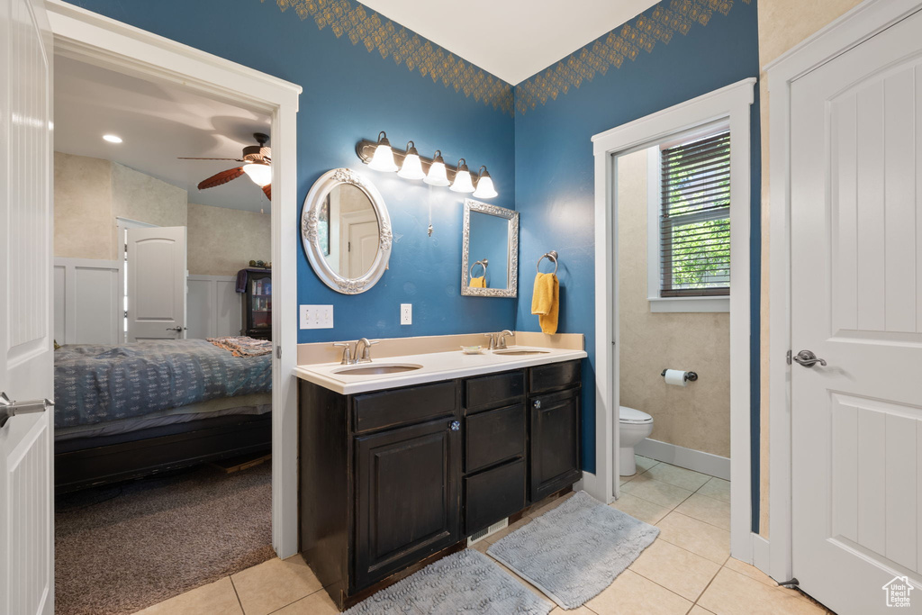 Ensuite bathroom featuring tile patterned floors, double vanity, ceiling fan, and wainscoting